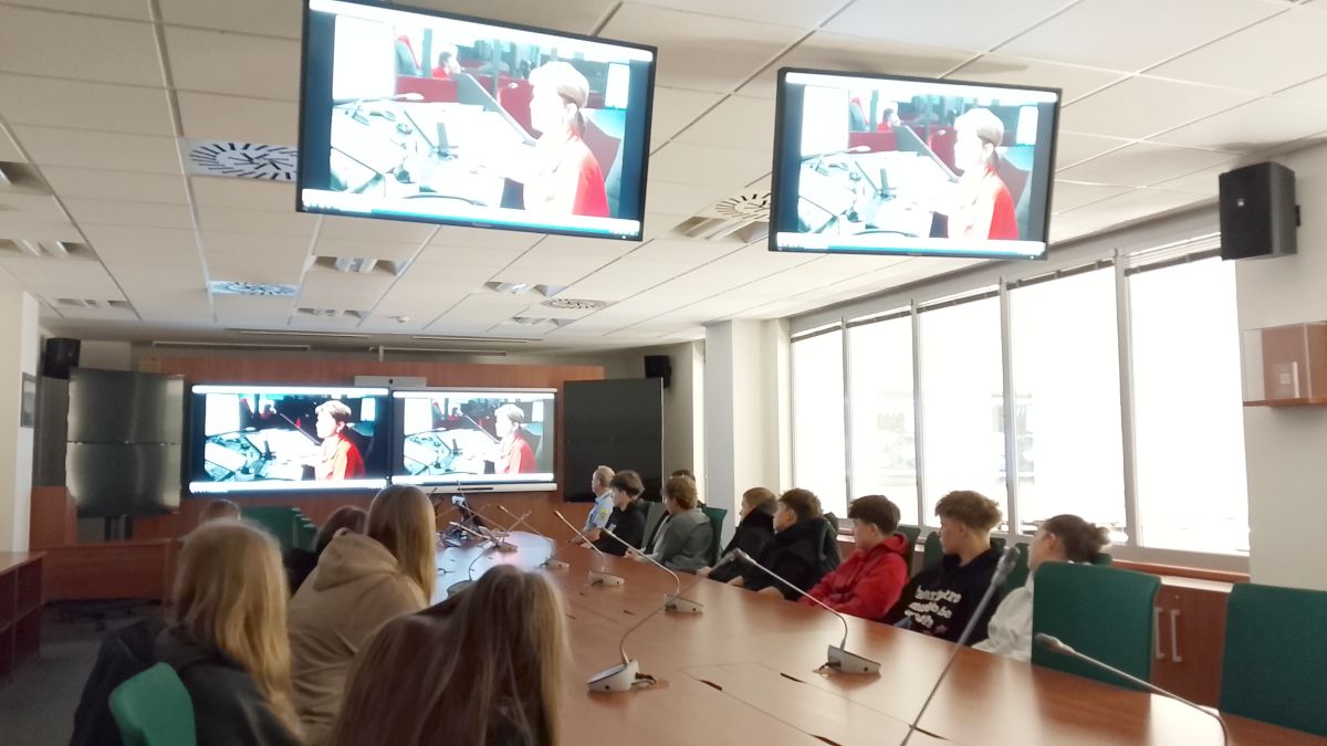 Žáci sedí u konferenčního stolu v zasedací místnosti a sledují na obrazovkách přenos soudního jednání.