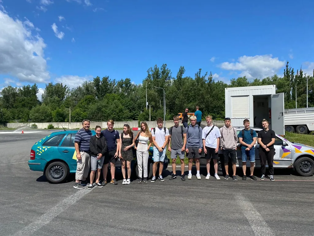 Skupina žáků a učitelů pózuje před vozidly autoškoly na zkušební dráze.