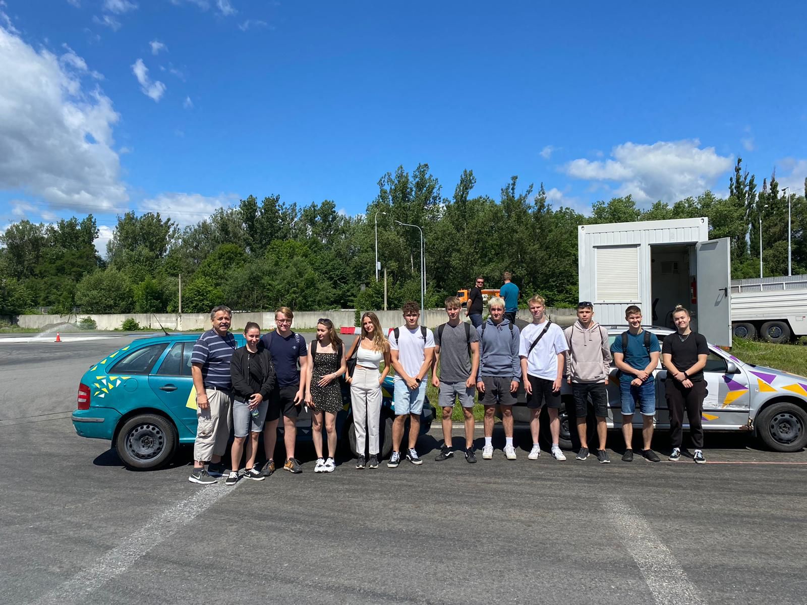 Skupina žáků a učitelů pózuje před vozidly autoškoly na zkušební dráze.