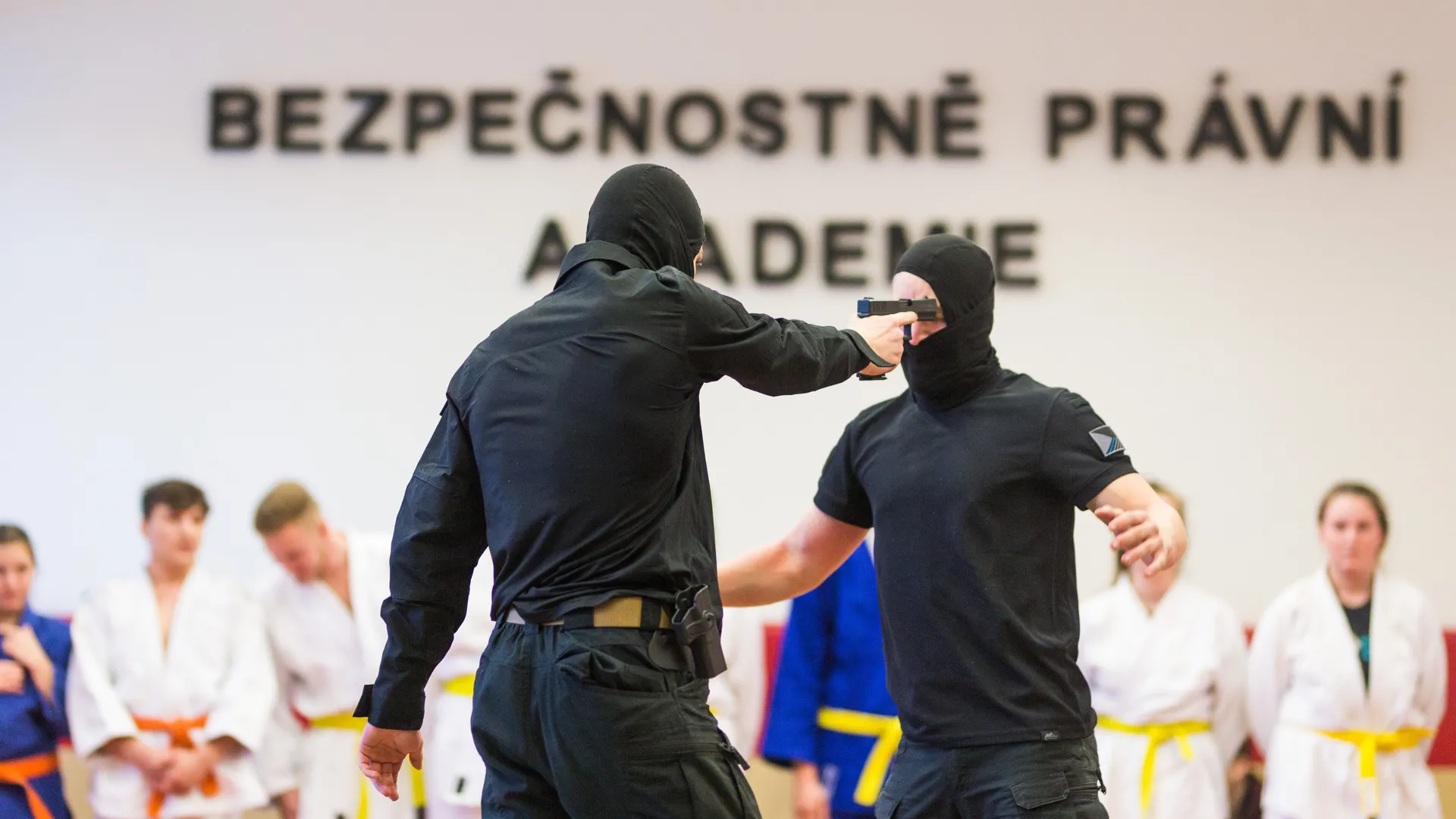 Dva žáci v černém oblečení a s kuklami nacvičuje obranu proti útoku střelnou zbraní. Odehrává se na tatami, v pozadí další žáci v kimonech.